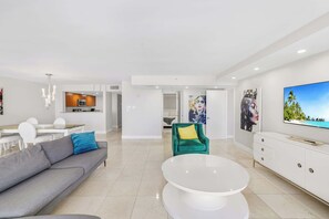 Una sala moderna con un sofá, una mesa redonda de centro, un sillón color turquesa, un televisor y una cocina al fondo.
