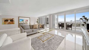 A modern living room with a sofa, coffee table, and a view of the city through large windows. - Central Long Stay Base | Gym & Balcony | StayCozy (Miami)