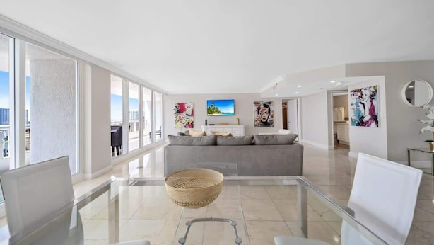 A spacious living room with a large window, a grey sofa, and a glass coffee table. - Central Long Stay Base | Gym & Balcony | StayCozy (Miami)