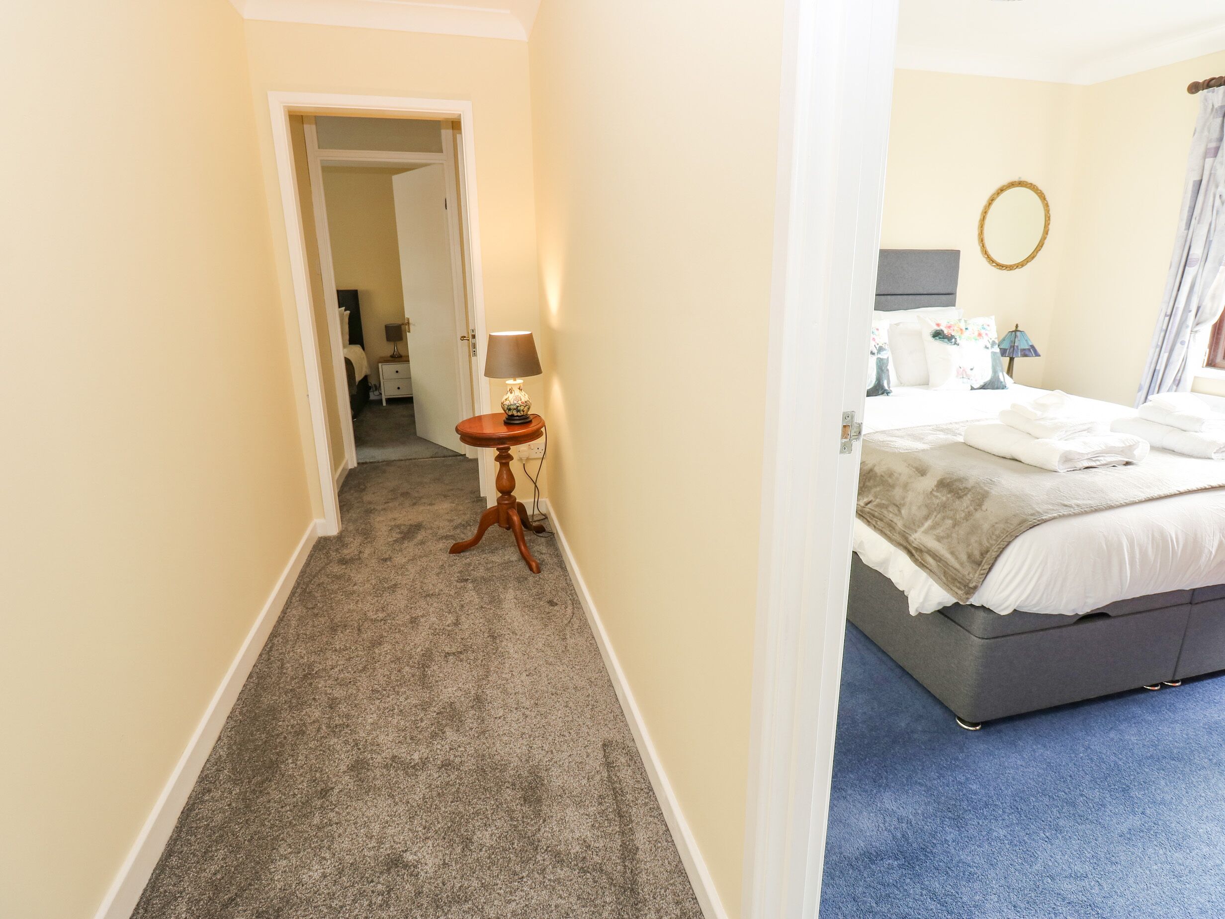 A hotel room with a bed, a small table with a lamp, a mirror, and a door leading to another room.