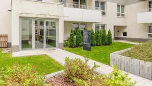 Een modern gebouw met een glazen ingang, een bord met daarop 'K1.7' en een verzorgde tuin.