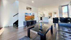 Una sala de estar moderna con un televisor de pantalla plana, una mesa de centro de vidrio y un sofá. Se ve una escalera que sube a otro nivel.