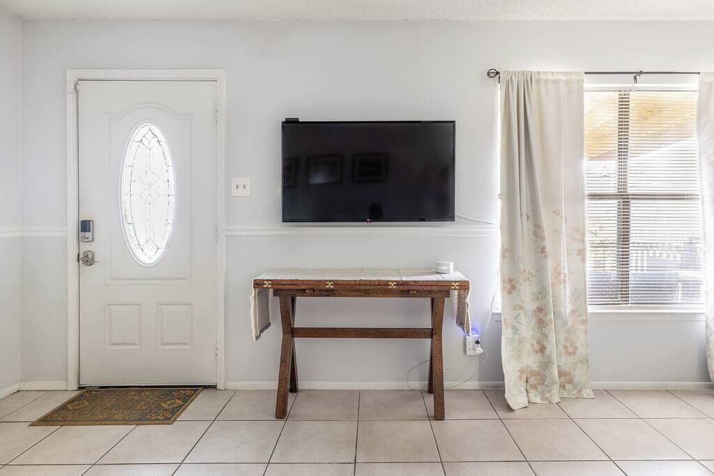 A room with a white door, a flat-screen TV mounted on the wall, a wooden table with a white cloth, and a window with blinds.