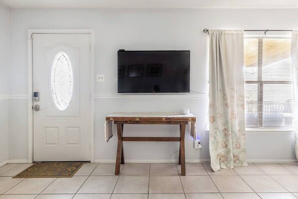 Una habitación con una puerta blanca, un televisor de pantalla plana montado en la pared, una mesa de madera con mantel blanco y una ventana con persianas.