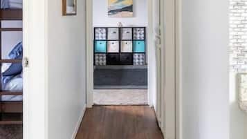 Een gang met houten vloer, een bed met blauw beddengoed, een lijst met afbeelding, een plank met opbergkisten, en een wandlamp.