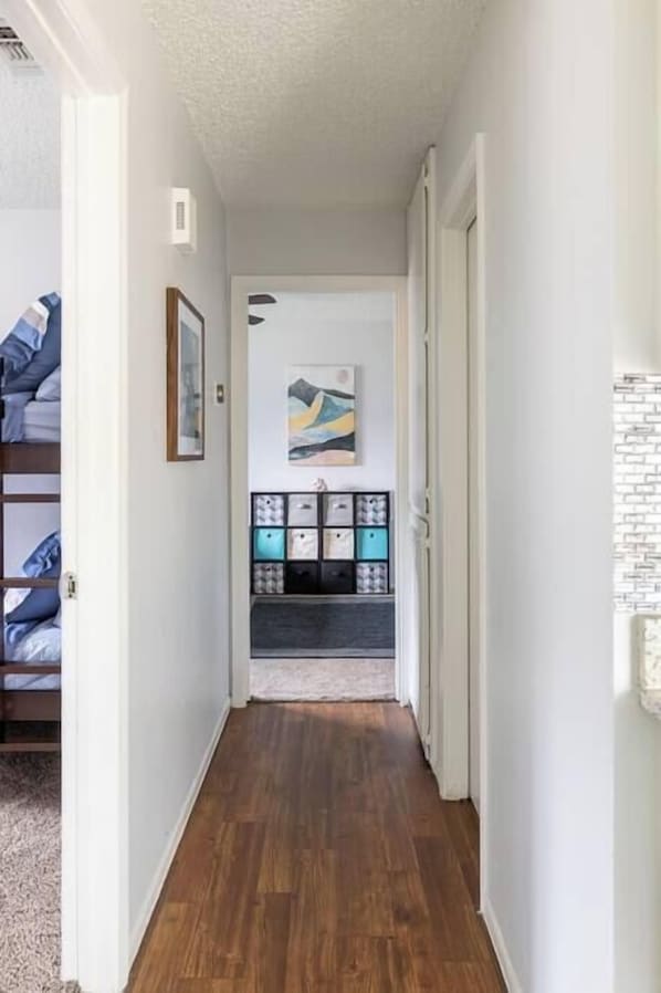 A hallway with wooden flooring, a bed with blue bedding, a framed picture, a shelf with storage boxes, and a wall-mounted light fixture.
