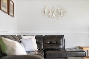 A living room with a dark leather sofa, a white wall, and a decorative leaf wall art piece.