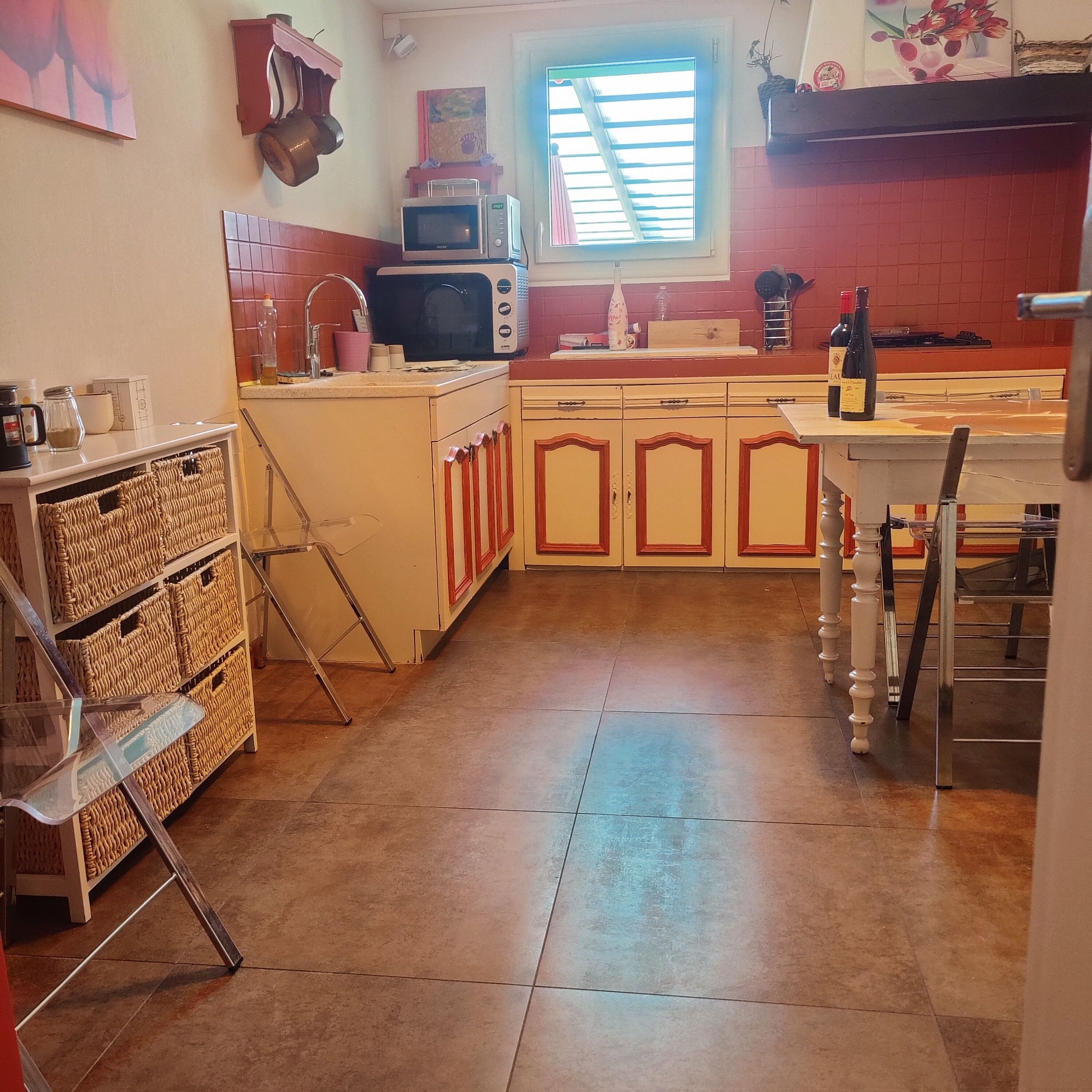 A kitchen with a microwave, sink, and cabinets. There is a window with blinds, a vase with flowers, and a bottle of wine on the counter.