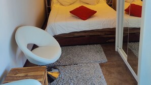A bedroom with a wooden headboard, a bed with white bedding and red pillows, a white chair, a small wooden table, and a mirror.