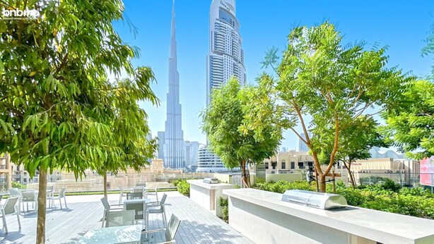 An outdoor seating area with tables and chairs, surrounded by greenery, overlooking a cityscape with a prominent skyscraper. - bnbme | Burj Royale | 1 BDR (Dubai)