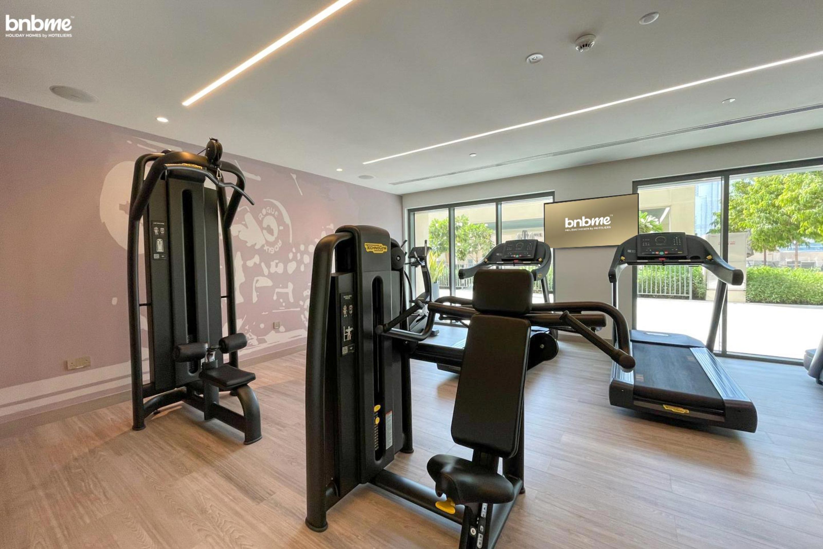 Une salle de sport équipée de matériel d’exercice, d’un tapis de course et de grandes fenêtres donnant sur l’extérieur.