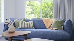 Una sala de estar moderna con un sofá azul, cojines a rayas, una manta color beige y un jarrón con flores secas sobre una mesita de madera.