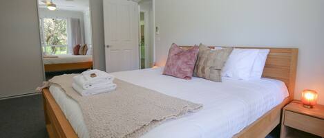 A neatly made bed with a white comforter, a beige throw blanket, and a stack of white towels. There is a wooden headboard, a bedside table with a lamp, and a mirror on the wall.