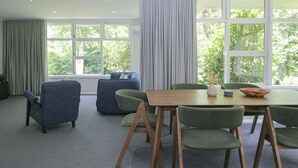 Un comedor moderno con una mesa de madera, sillas tapizadas en tonos verde y azul, y amplios ventanales que ofrecen vistas a zonas verdes.