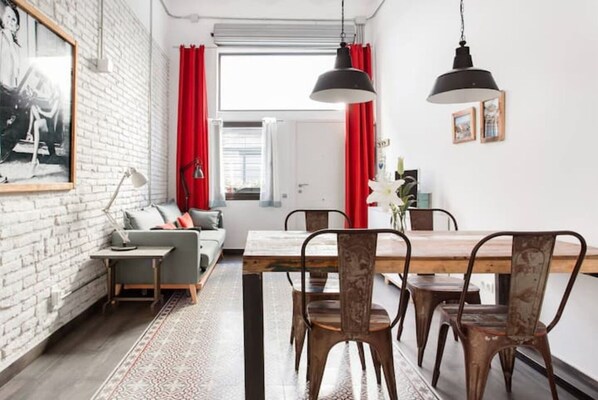 A modern living room with a sofa, a dining table with chairs, a patterned rug, and a large window with red curtains.