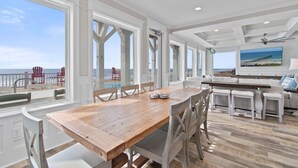A spacious room with a large wooden table and white chairs, a bar area with stools, and large windows offering a view of the beach. - Experience luxury at '7 Day Weekend,' an oceanfront 7-bedroom retreat in Oak Island. (Oak Island)