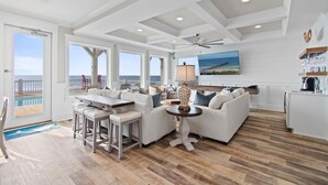A spacious living area with a large window overlooking the ocean, a ceiling fan, and a kitchen area with a view of the sea. - Experience luxury at '7 Day Weekend,' an oceanfront 7-bedroom retreat in Oak Island. (Oak Island)