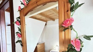 Espejo de marco de madera con rosas rosadas en la pared, reflejando una habitación con cama, cómoda y una mesita.