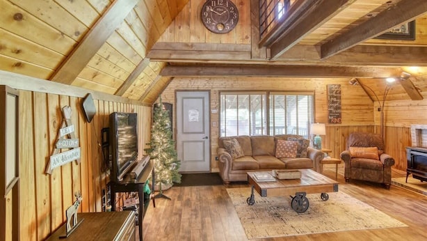 A cozy cabin interior with a wooden ceiling, a living room with a sofa and a coffee table, a fireplace, and a Christmas tree.