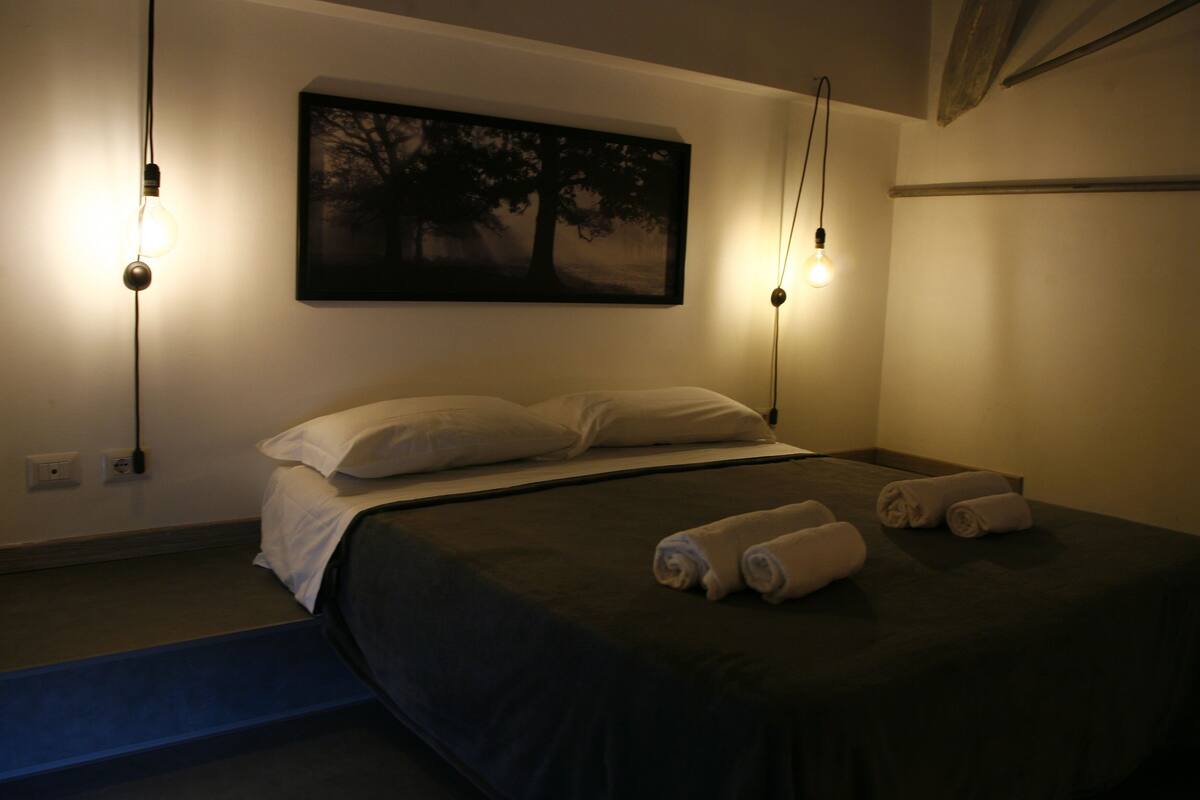A bedroom with a bed, two bedside lamps, a painting of a tree, and rolled towels on the bed.