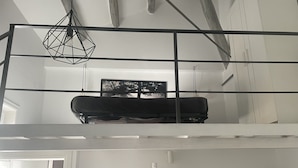 Ein Loft-Zimmer mit einem an einem Metallgestell hängenden Bett, einem an der Wand befestigten Fernseher und einem Regal mit diversen Gegenständen.