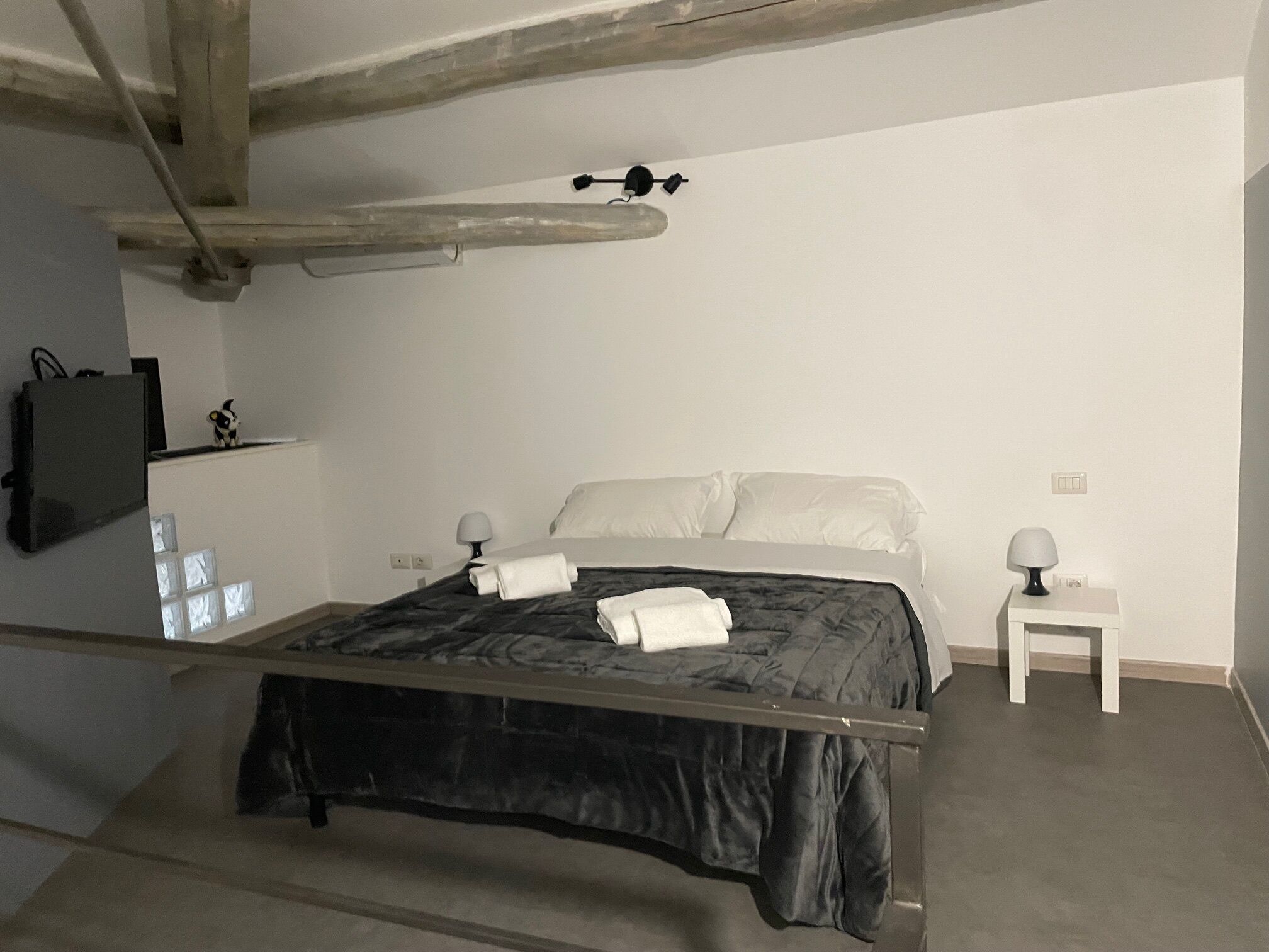 A bedroom with a bed, two bedside tables, a wall-mounted TV, and a window with blinds.