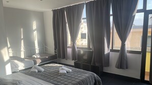 Ein modernes Hotelzimmer mit einem großen Bett, zwei kleineren Betten, einem Fernseher und einem Balkon mit Vorhängen.