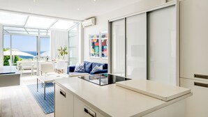 A modern kitchen with a large island, a dining area with a table and chairs, a sofa, and a view of the ocean. - Compact Camps Bay Holiday Apartment With Private Pool Sea Escape (Cape Town)