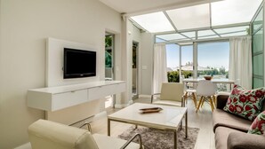 A modern living room with a flat-screen TV, white furniture, a glass-enclosed dining area, and a view of the outdoors. - Compact Camps Bay Holiday Apartment With Private Pool Sea Escape (Cape Town)