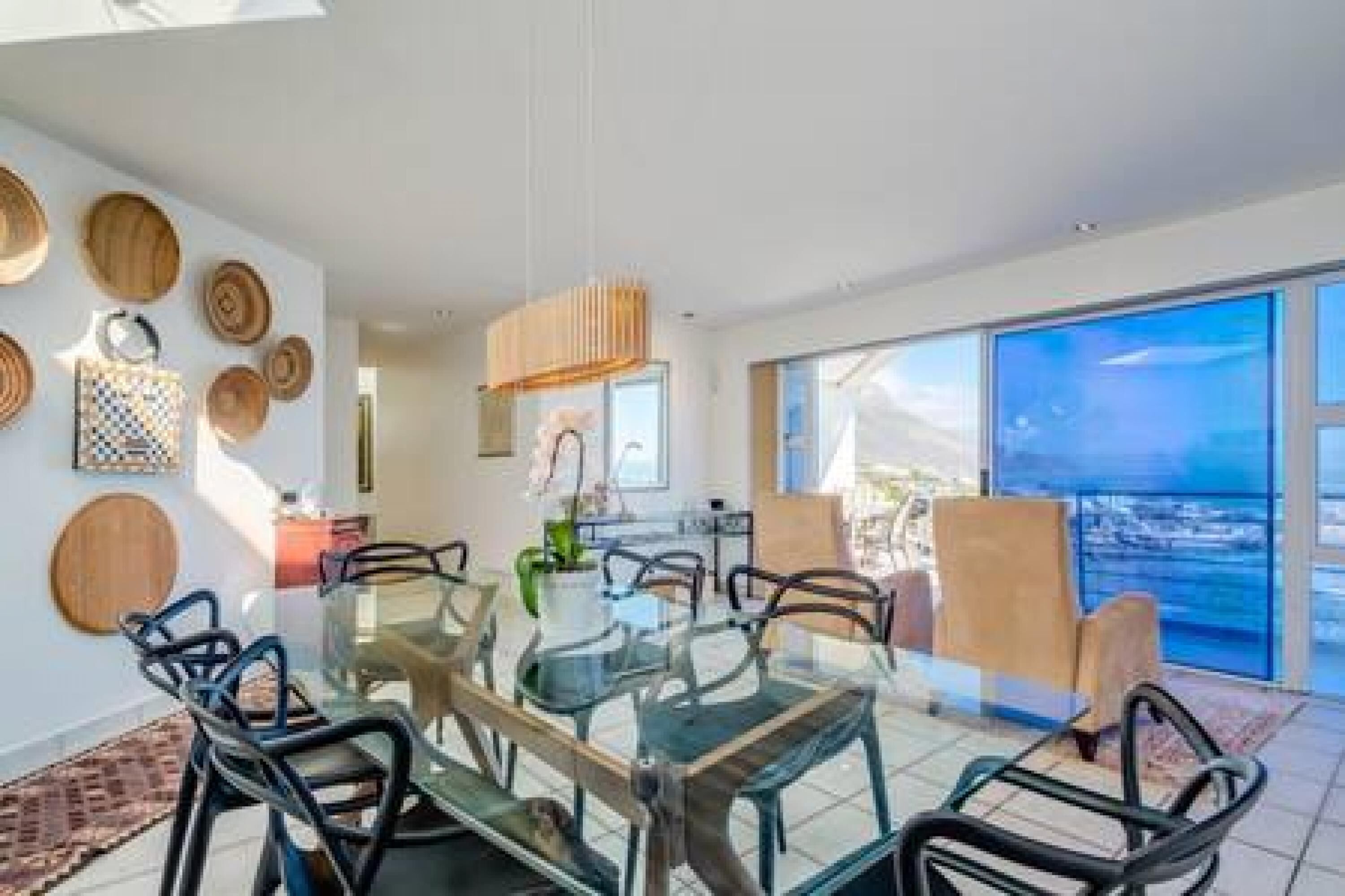 A modern dining area with a glass table and metal chairs, surrounded by large windows with a view of the ocean.