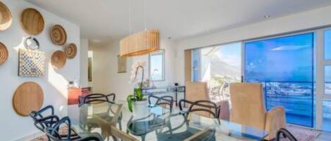 A modern dining area with a glass table and metal chairs, surrounded by large windows with a view of the ocean.