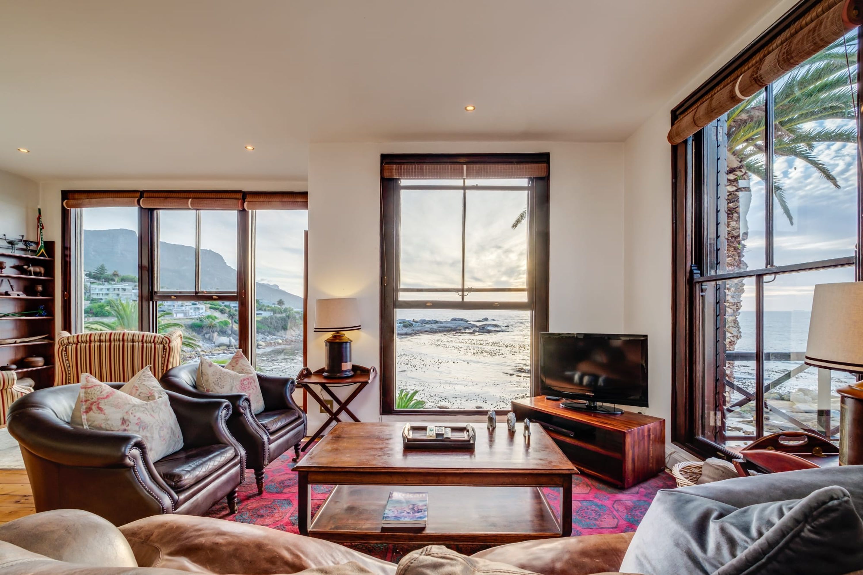 Amplia sala de estar con un gran ventanal que ofrece vistas a la playa y las montañas. Hay un sofá de cuero, una mesa de centro y un televisor.