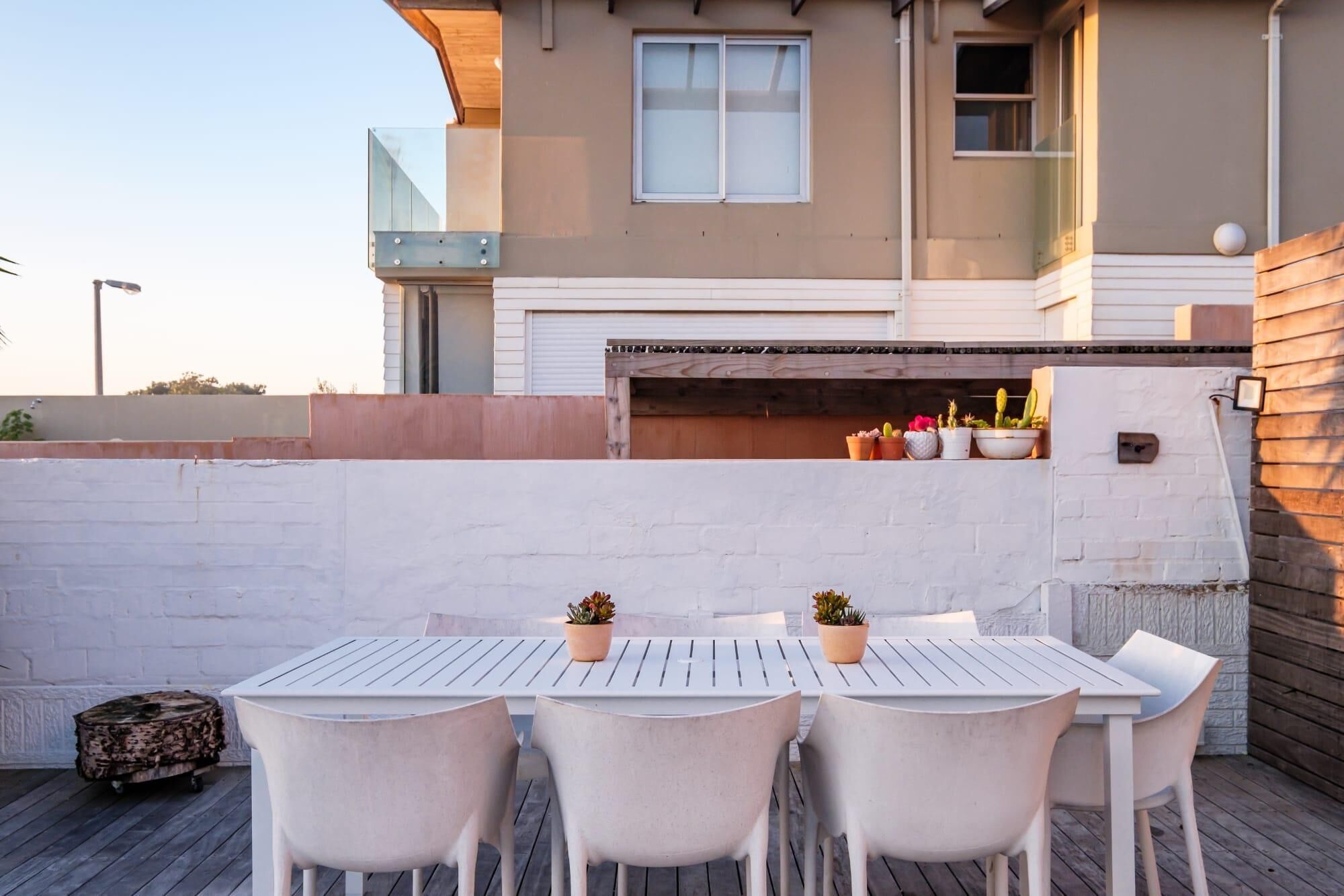 Un patio con una mesa y sillas blancas, una terraza de madera y un edificio con ventanal y un pequeño balcón con plantas en macetas.