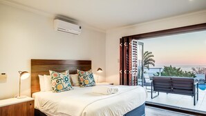 Une chambre avec un grand lit, une table de chevet, une lampe et une vue sur l’océan par une porte coulissante.
