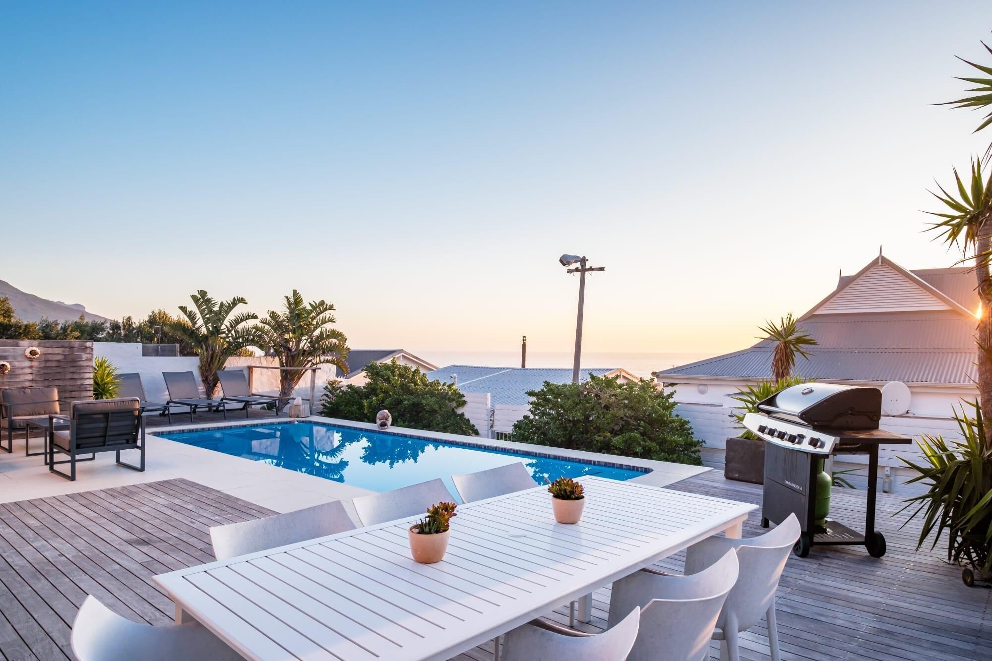 A rooftop pool area with a barbecue grill, lounge chairs, and a table with potted plants.