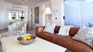 A modern living room with a brown leather sofa, a white coffee table with a bowl of fruit, and a large window with a view. - Holiday Apartment w Sea Views Pool CBT Suite (Cape Town)