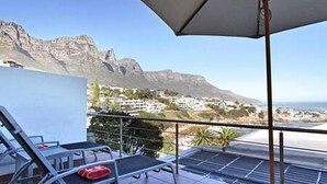 A balcony with a table, chairs, and an umbrella, overlooking a mountainous landscape and a coastal area. - Holiday Apartment w Sea Views Pool CBT Suite (Cape Town)