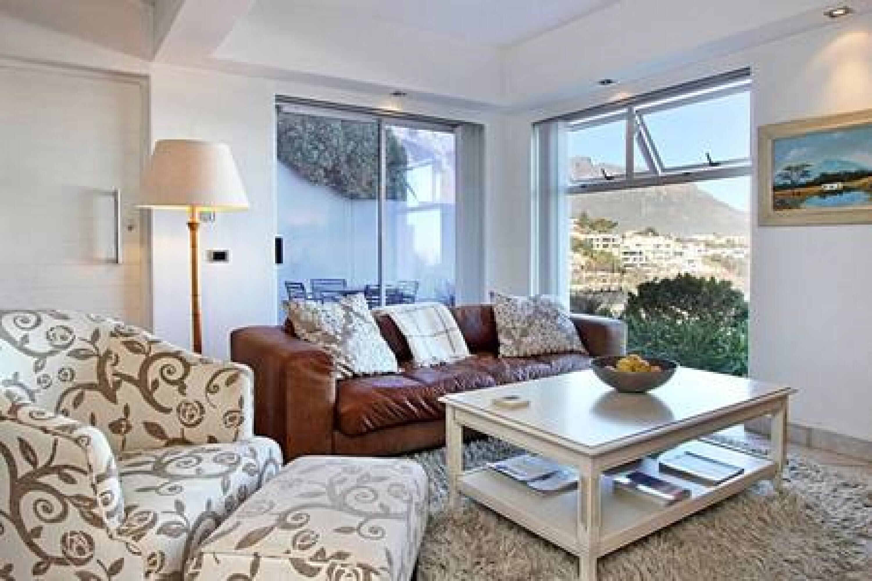 Ein modernes Wohnzimmer mit einem braunen Ledersofa, einem gemusterten Sessel und einem Couchtisch. Ein großes Fenster bietet Blick auf die Stadt.