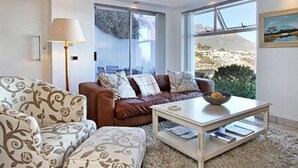 A modern living room with a brown leather sofa, patterned armchair, and a coffee table. A large window offers a view of a cityscape. - Holiday Apartment w Sea Views Pool CBT Suite (Cape Town)