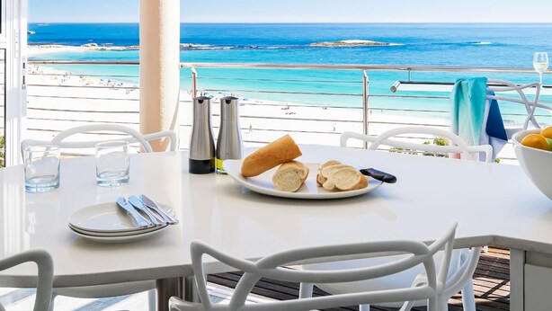 A table with a view of the beach and ocean, a plate with bread, and a glass of water. - Beautiful Holiday Apartment Overlooking the Beach Modoco (Cape Town)
