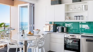 A modern kitchen with white cabinets, a green backsplash, and stainless steel appliances. There is a dining area with white chairs and a table, and a view of the ocean through a large window. - Beautiful Holiday Apartment Overlooking the Beach Modoco (Cape Town)