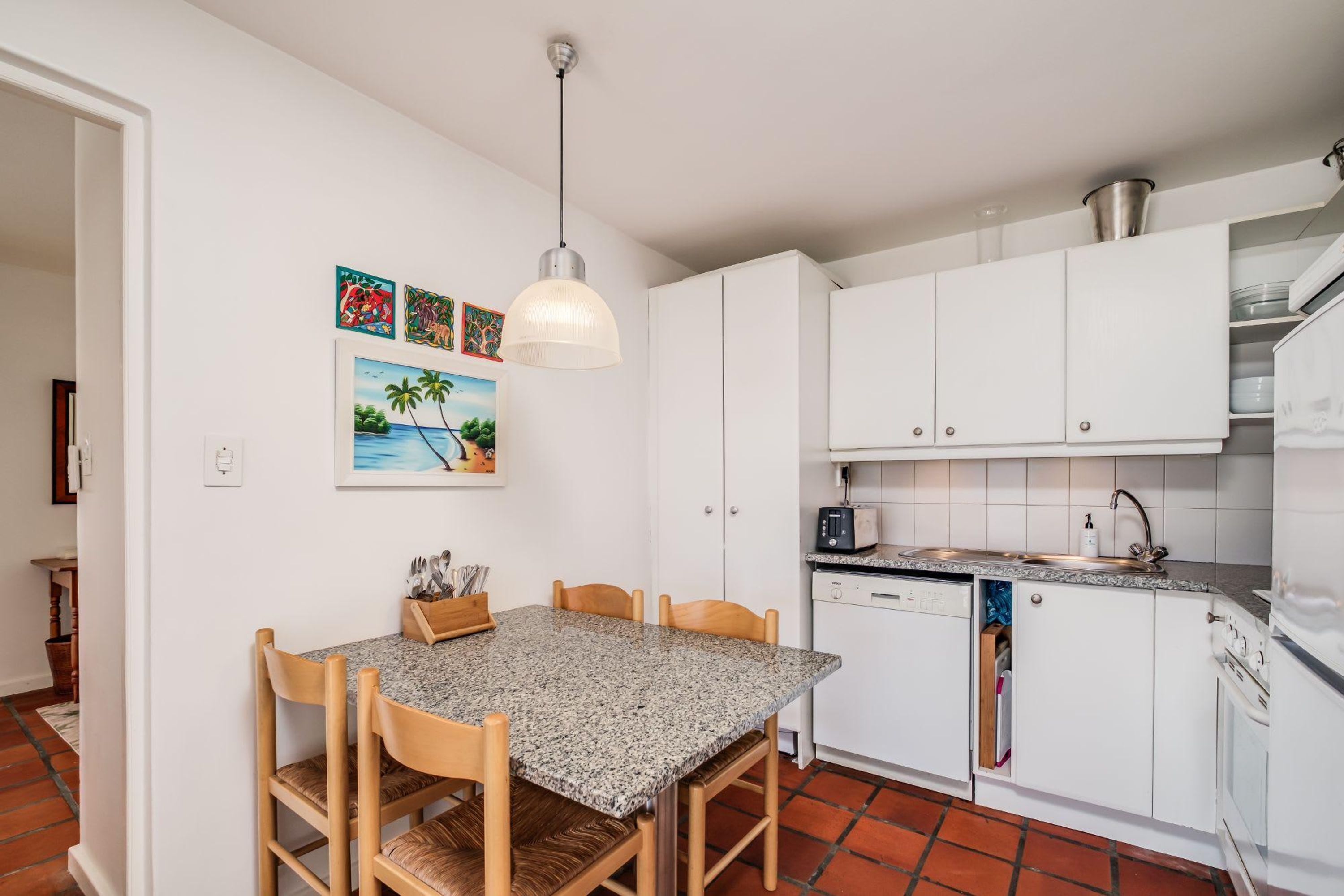Cozinha compacta com armários brancos, bancada de granito, lava-louças embutida e área de refeições com mesa e cadeiras.