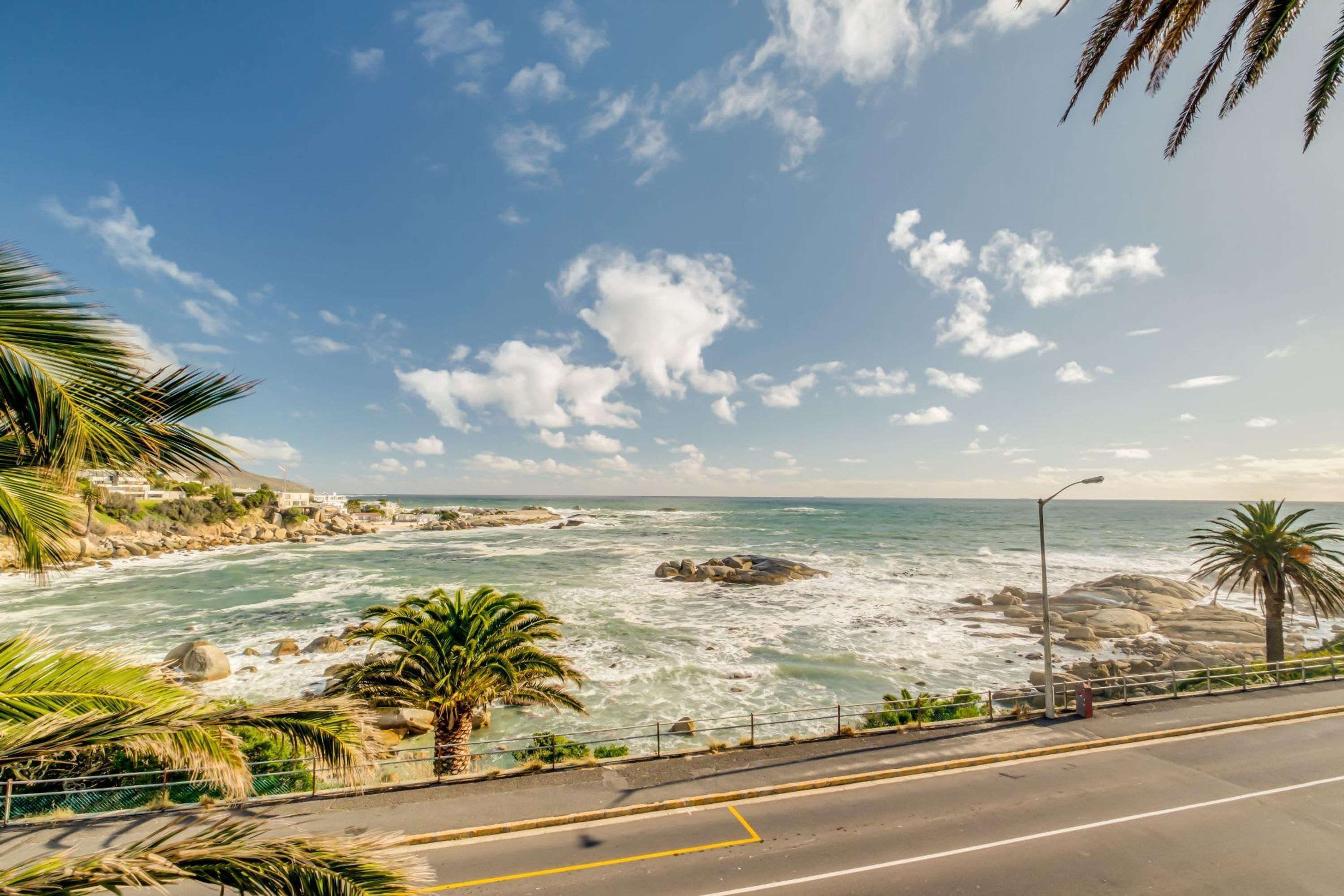 A coastal road with a view of the ocean, palm trees, and rocky outcrops.