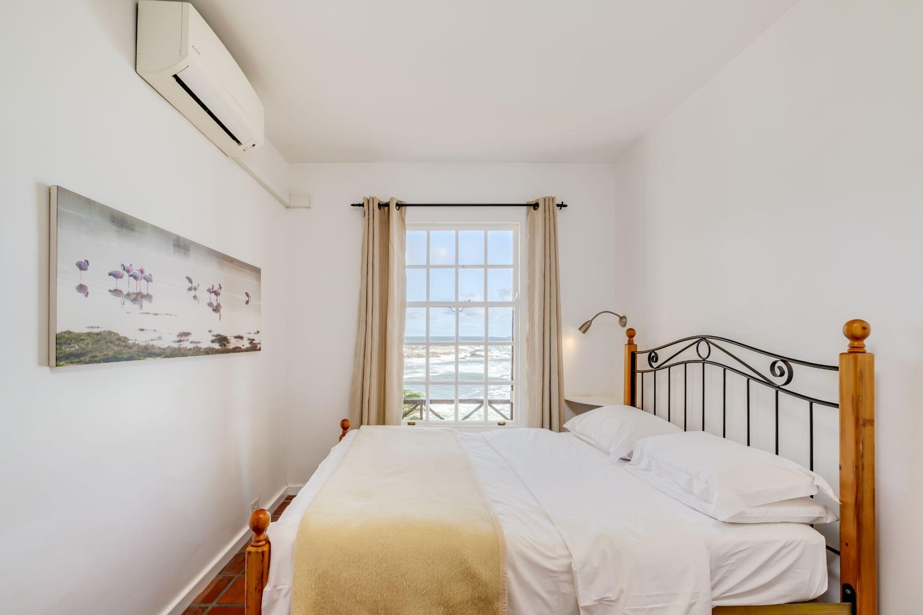 A bedroom with a bed, a window with curtains, an air conditioning unit, and a painting on the wall.
