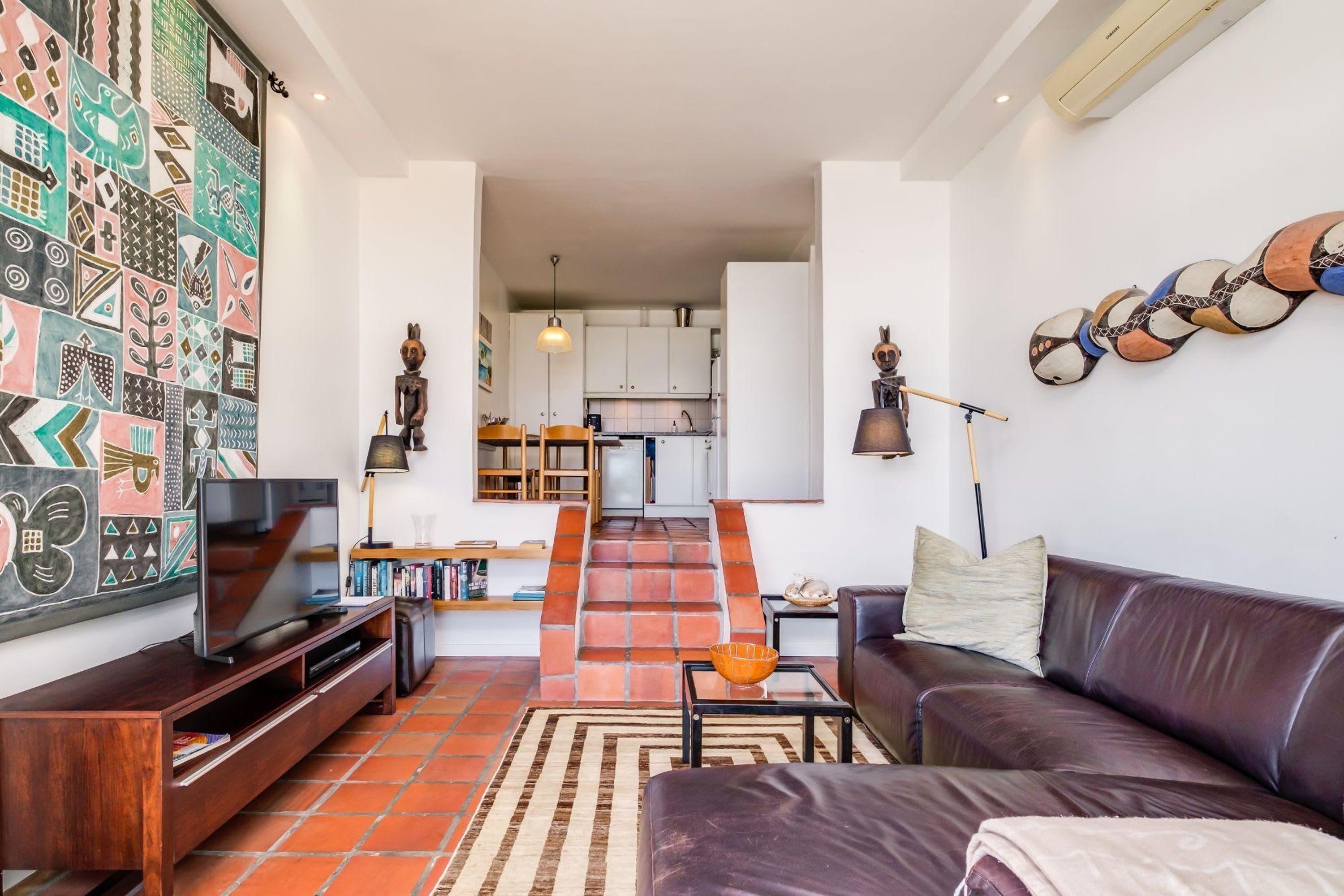 Ein modernes Wohnzimmer mit Fernseher, Sofa und Couchtisch. Eine Treppe führt zu einer Küche mit weißen Schränken und einer Pendelleuchte.