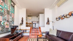 A modern living room with a television, sofa, and coffee table. A staircase leads to a kitchen with white cabinets and a hanging light fixture. - Camps Bay Holiday Apartment w Private Pool CBT Palm Suite (Cape Town)