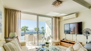 Ein modernes Wohnzimmer mit einem Glastisch, einem beigefarbenen Sofa und einem Flachbildfernseher. Ein großes Fenster bietet Blick auf den Ozean und Palmen.