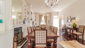 A spacious dining room with a long wooden table, surrounded by chairs with patterned cushions. A fireplace with decorative items is on the left, and a chandelier hangs from the ceiling. - Lovely Holiday Home w Private Courtyard Six Selbourne (Cape Town)