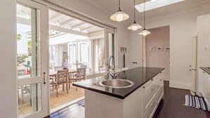 A modern kitchen with a large island, a double sink, pendant lighting, and a view to an outdoor dining area. - Lovely Holiday Home w Private Courtyard Six Selbourne (Cape Town)