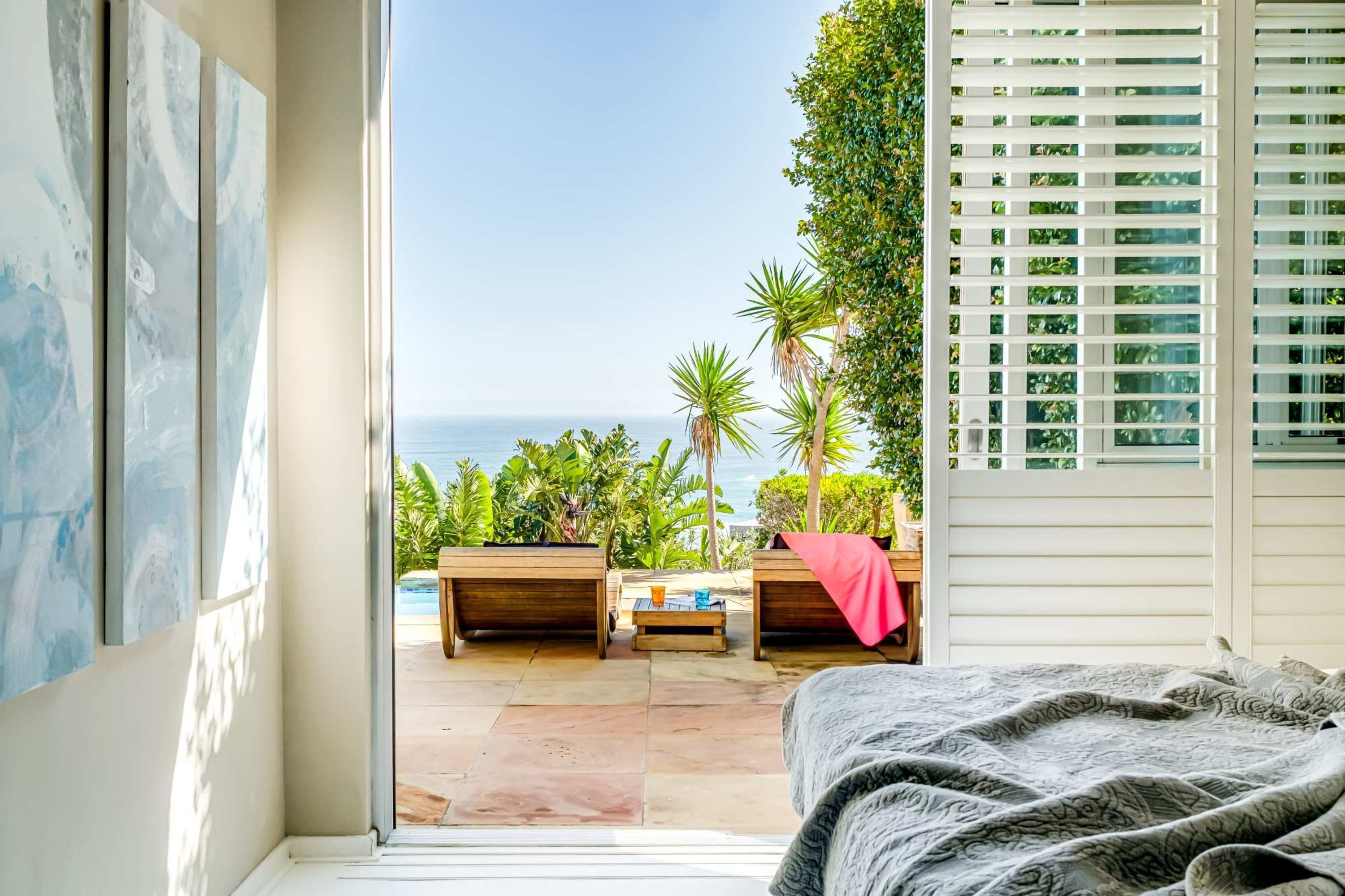 A bedroom with a view of the ocean, a bed, a window with blinds, and a small table with a red cloth.